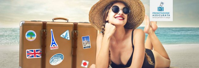 Donna con cappello sulla spiaggia accanto a una valigia.