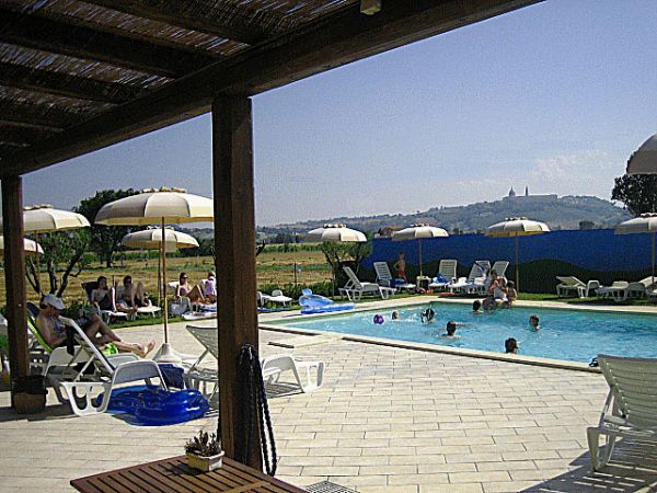 Piscina all'aperto con lettini e ombrelloni, persone che nuotano e panorama collinare sullo sfondo.
