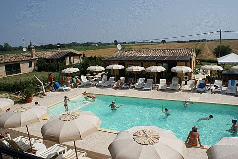 Piscina all'aperto con ombrelloni bianchi, lettini e persone che nuotano.