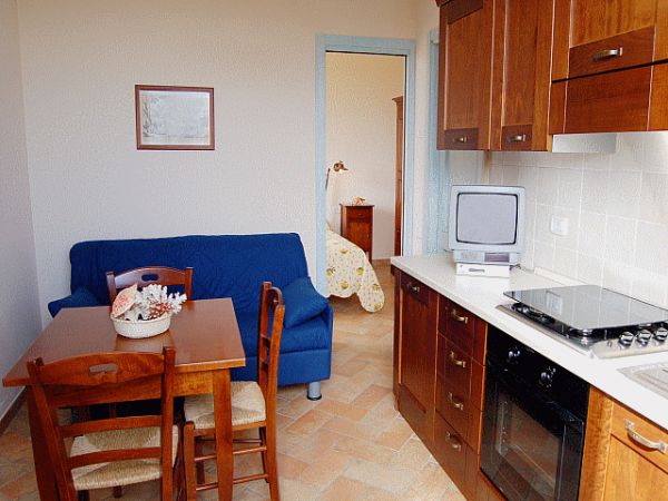 Cucina con tavolo in legno, sedie, divano blu e televisione. In fondo, una camera da letto.