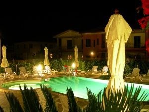 Piscina illuminata di notte con sdraio e ombrelloni chiusi.