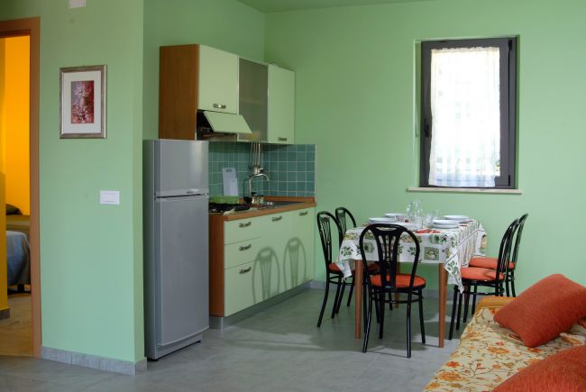 Cucina moderna con pareti verdi, tavolo apparecchiato, sedie nere e divano arancione.