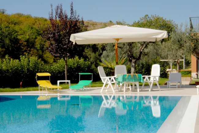Piscina con lettini colorati e un grande ombrellone bianco, circondata da alberi e cespugli.