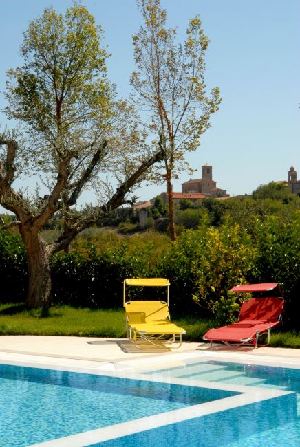 Una piscina con due sdraio colorate, alberi e un borgo sullo sfondo.