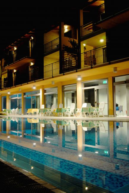 Piscina illuminata di notte con sedie a sdraio e palazzina a più piani sullo sfondo.