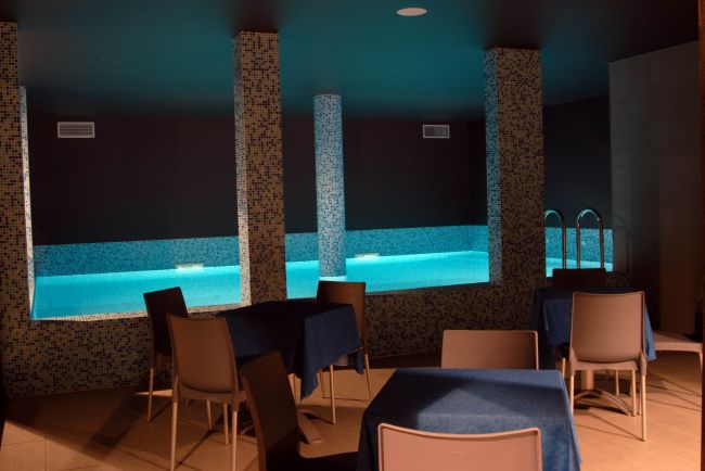 Piscina interna con illuminazione blu, tavoli e sedie intorno.