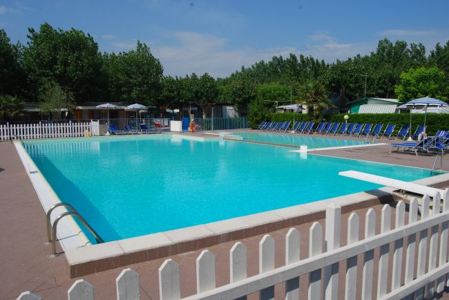 Piscina circondata da sdraio blu e recinzione bianca, con alberi sullo sfondo.
