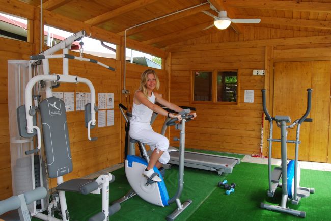 Donna su cyclette in una palestra di legno con attrezzi fitness e tappeto verde.