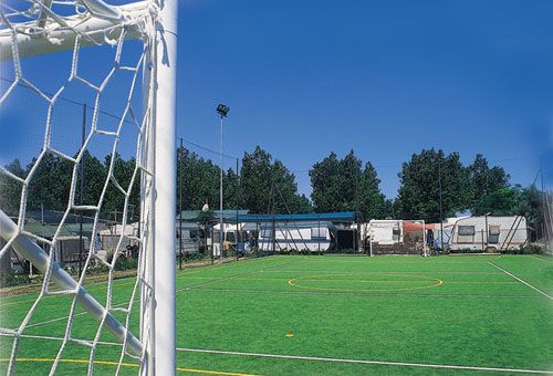 Campo da calcio in erba sintetica con rete in primo piano e camper dietro la recinzione.