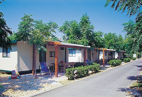 Esterno di un campeggio con bungalow, sedie a sdraio e ombra degli alberi.
