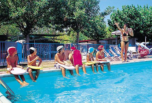 Bambini seduti al bordo di una piscina ascoltano un istruttore.
